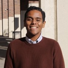 Jeffrey Barrera head shot standing in a brown sweater smiling outside of stadium